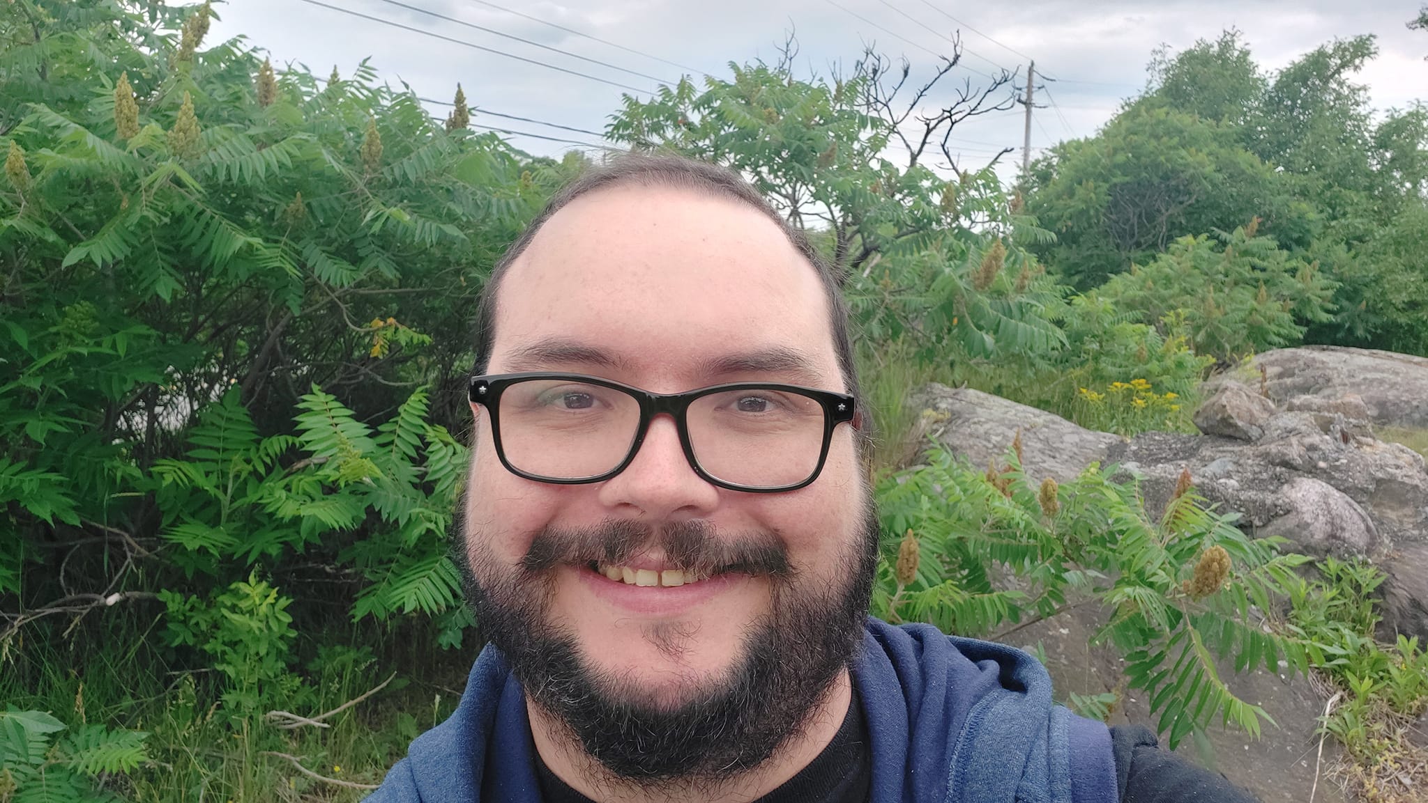 A man is dressed in a t-shirt and hooded sweater vest sits in front of some shrubs and Canadian shield bedrock